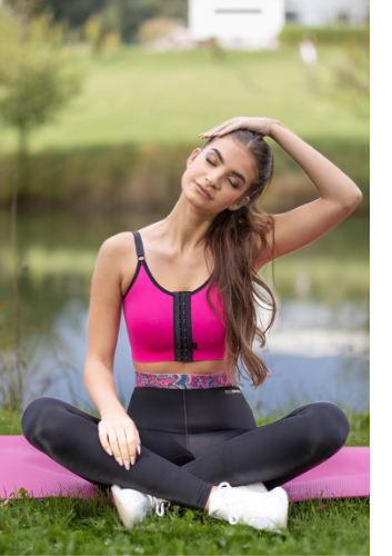 Eine Frau mit braunen Haaren sitzt auf einer pinken Yogamatte im Schneidersitz und trägt eine schwarze ACTIVE leggings mit buntem Bund und den PI relax Kompressions-BH von LIPOELASTIC in der Farbe pink.